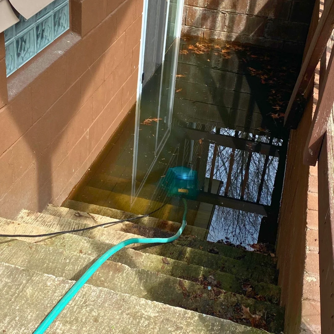 Outdoor stairwell flooded with water and a pump with hose removing the water near a glass door.