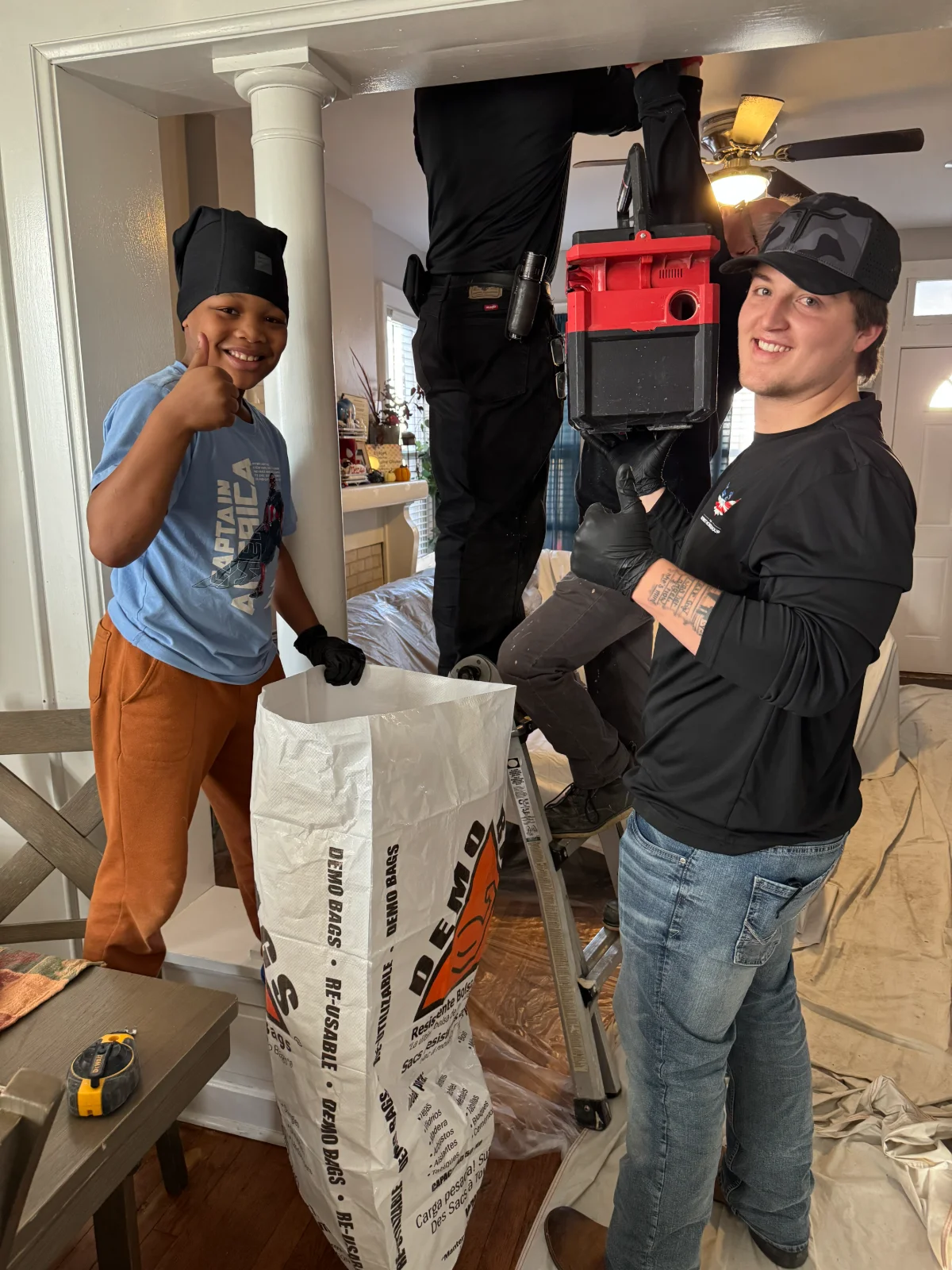 Three people doing home repair: one on ladder, one holding toolbox, boy giving thumbs up by a large bag.