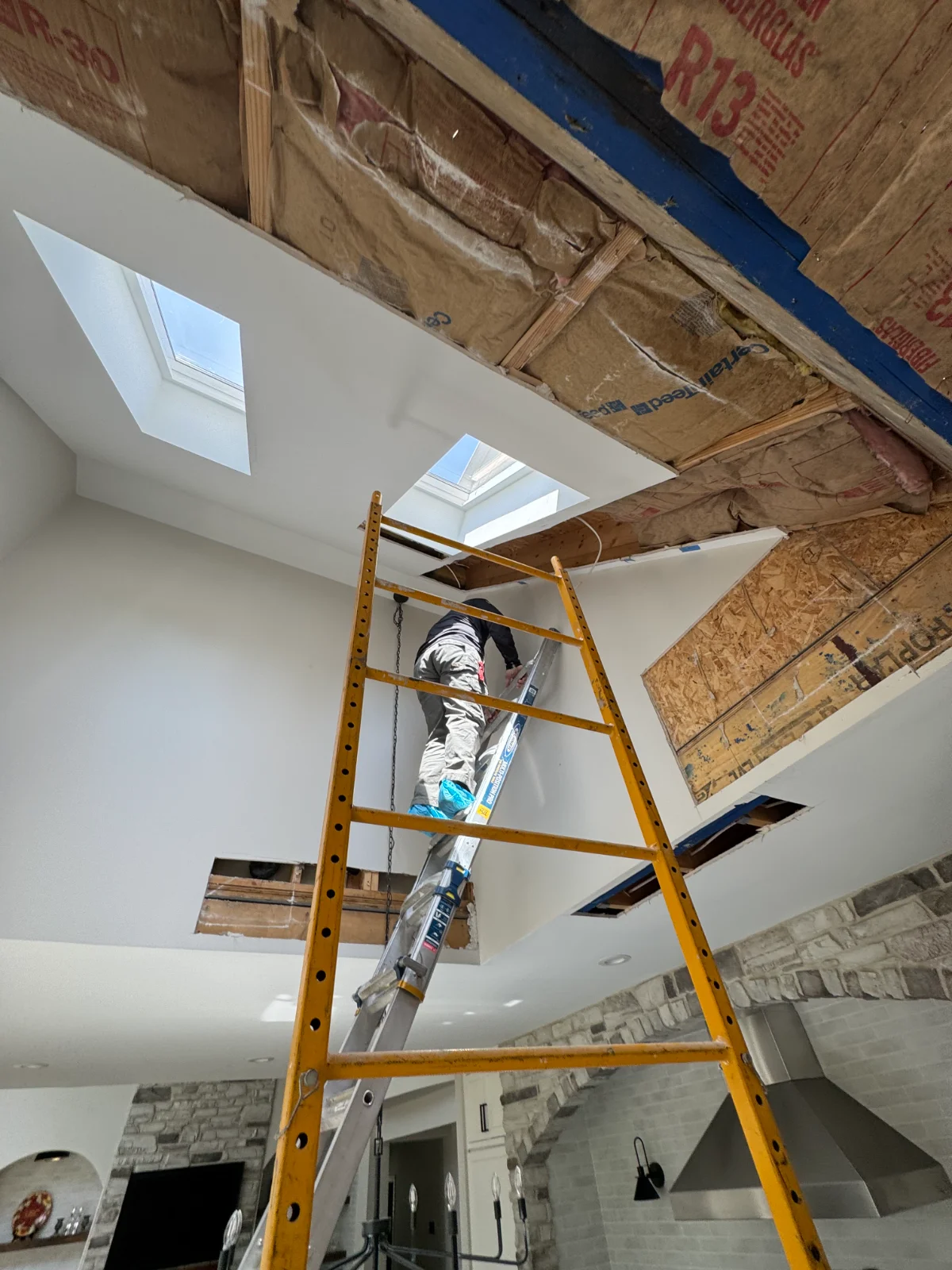 Worker on ladder repairing ceiling near skylights with exposed insulation and wooden beams.