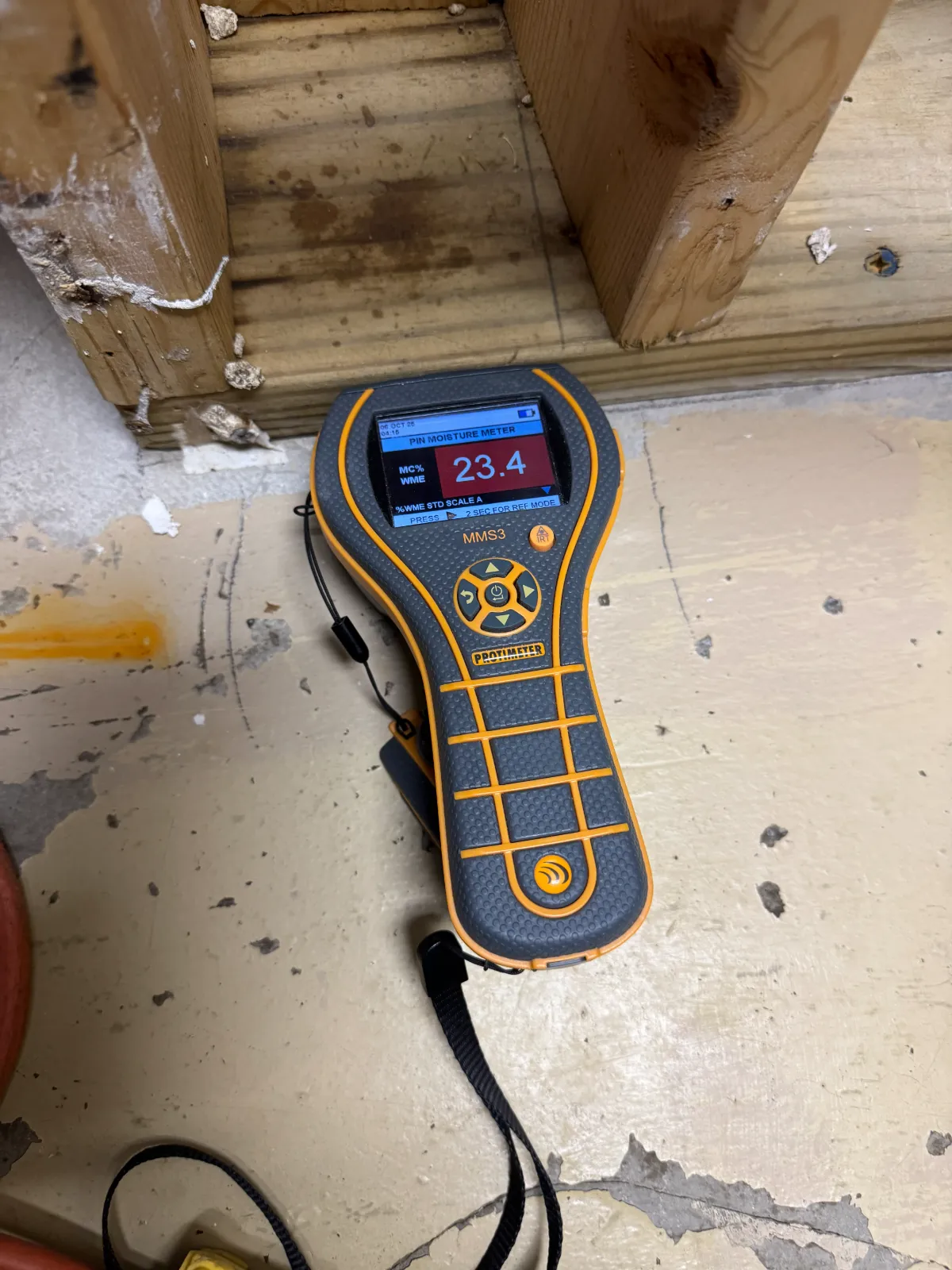 Handheld moisture meter on floor showing a reading of 23.4 percent near wooden beams.