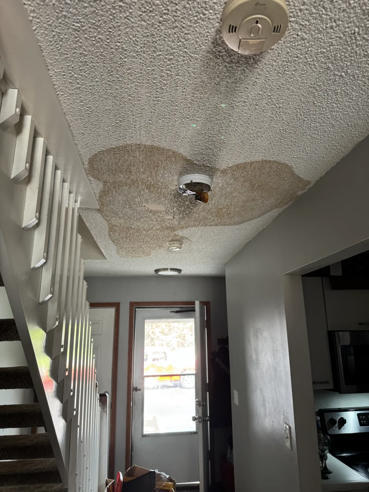 Hallway ceiling with large brown water stains and damaged smoke detector near an entrance door.