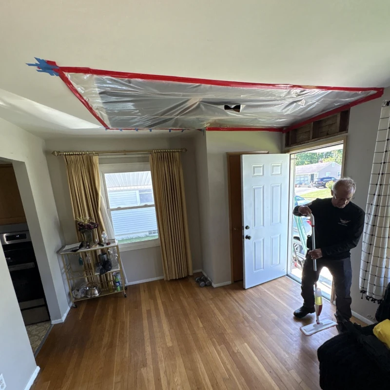 Person cleaning hardwood floor in a room with ceiling covered in taped plastic sheeting and open door.