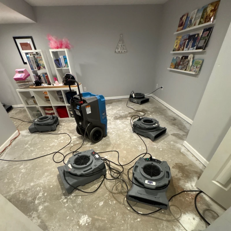 Basement room with industrial fans and dehumidifier on a wet, unfinished concrete floor for water damage repair.