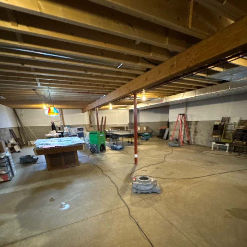 Basement with exposed ceiling beams, concrete floor, drying equipment, ladder, and scattered furniture.
