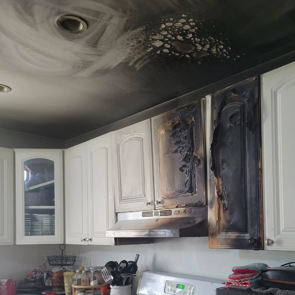 White kitchen cabinets and ceiling with severe black fire damage and soot above stove area.