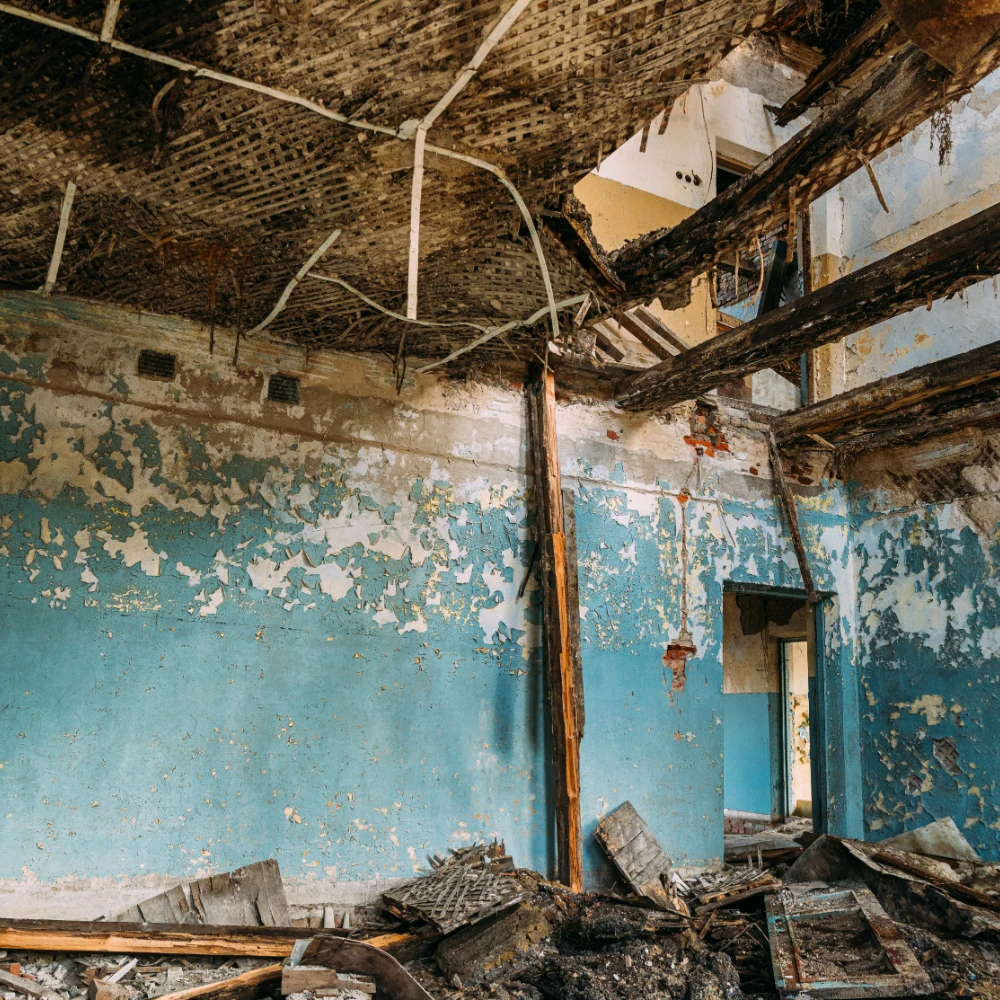 Room with heavy fire damage, charred beams, peeling blue paint, debris, and collapsed ceiling.