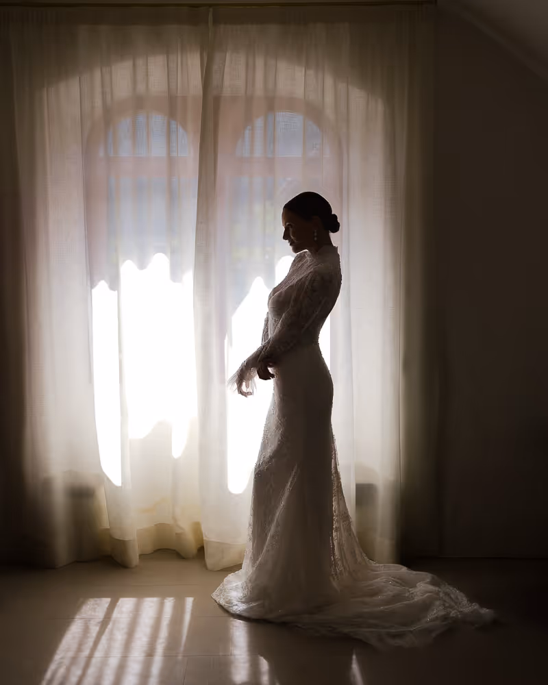 Bride adjusts her sleeves in elegant wedding gown in front of soft window light. 