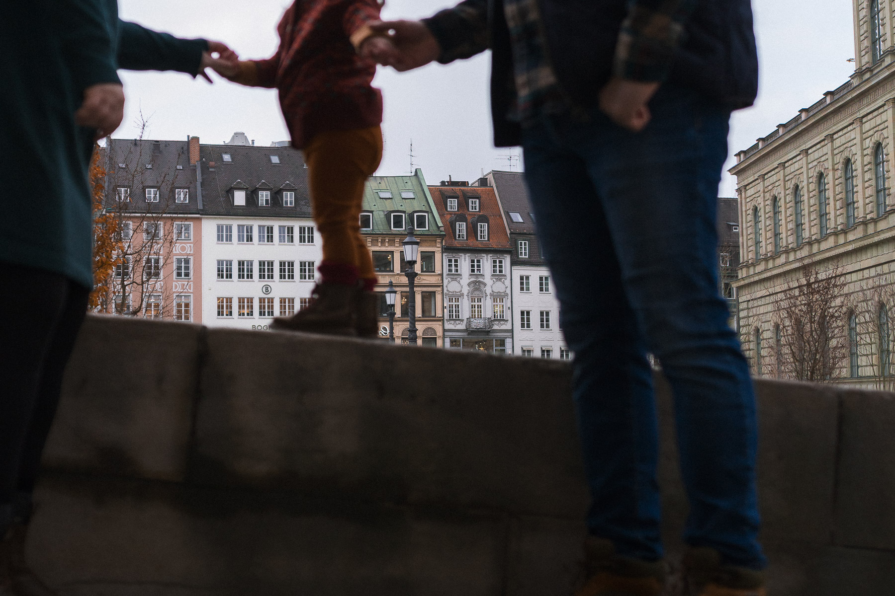 Little girl holding her parents hands, while walking across a stone railing with the Max-Joseph-Platz colourful architecture in the background.