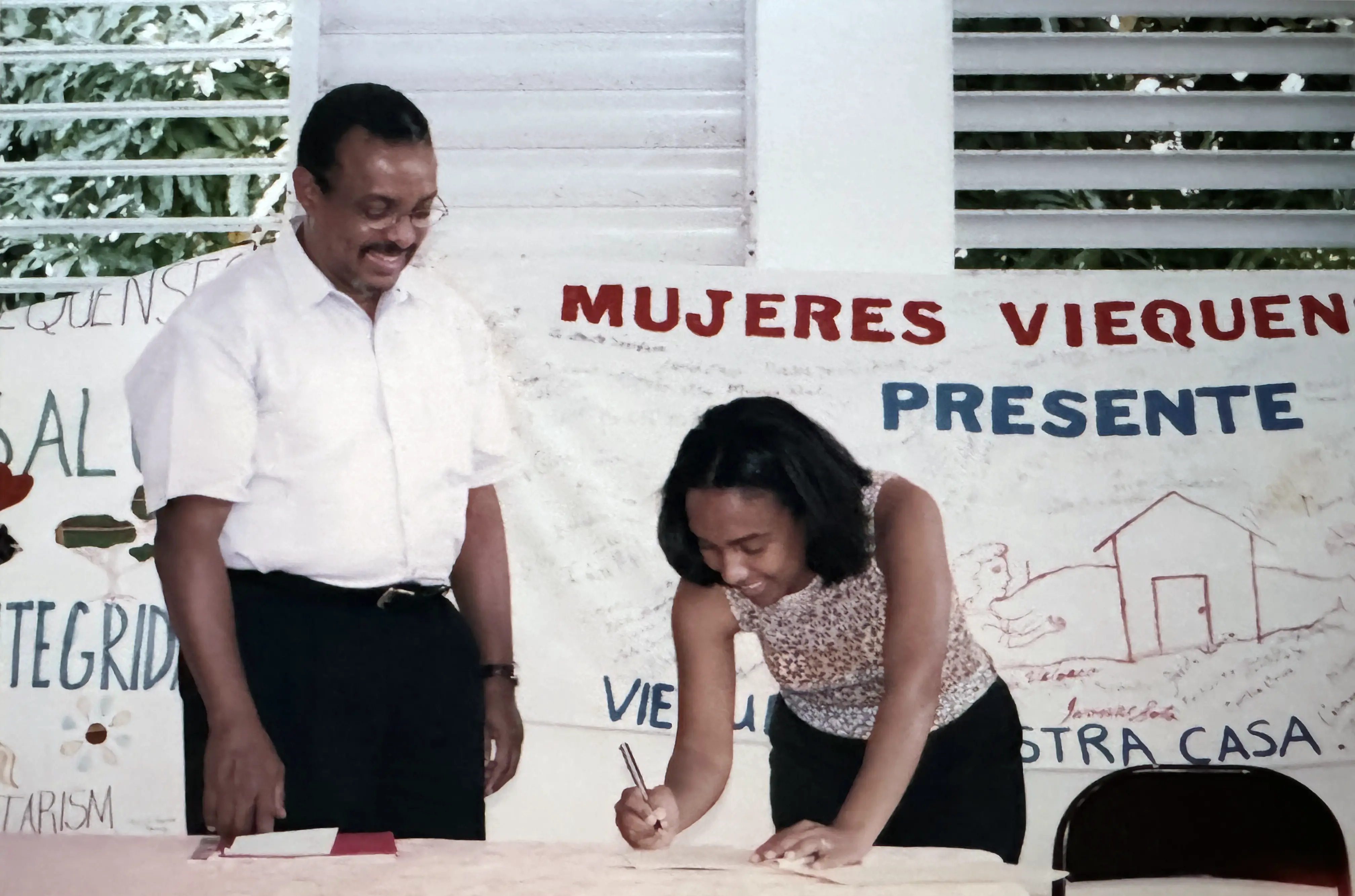 Judith Conde firmando proyecto con Nelson Colón Tarrats de Fundación Comunitaria frente a la primera pancarta de la Alianza de Mujeres Viequenses (2000, suministrada por la Alianza de Mujeres Viequenses).