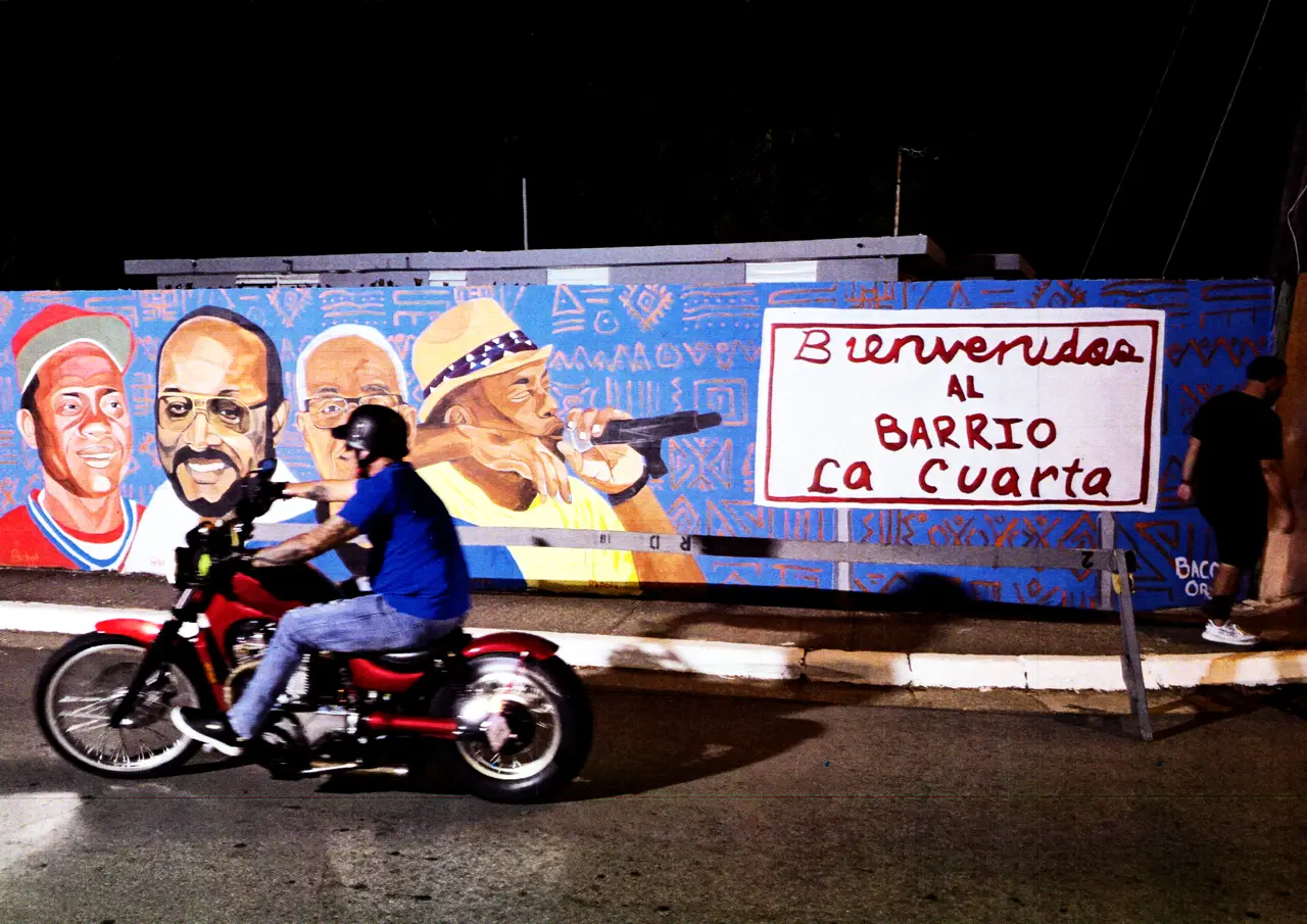 Motociclista frente al mural a la entrada del parque Barrio La Cuarta realizado por Jean “Baco” Ortiz en el 2024 con las figuras de: Juan Pachot Santiago, Rafael “Chago” Santiago Erans, José Rivera Guilbe y Ángel “Papote” Alvarado Aguilera.