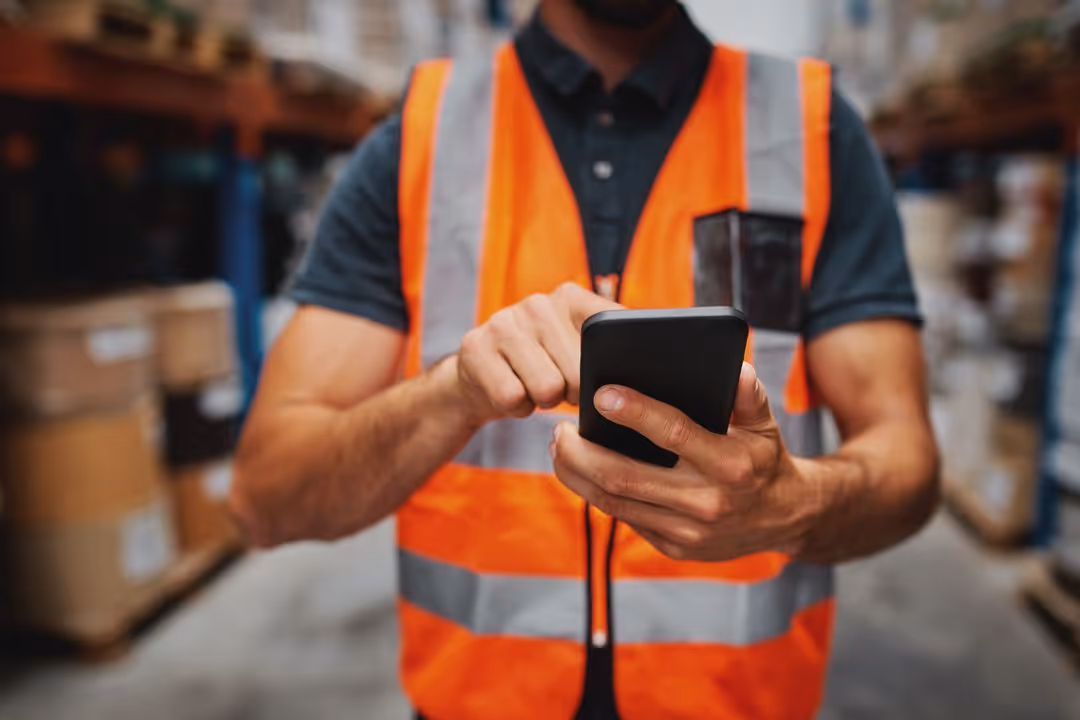 Warehouse worker in orange safety vest using a smartphone to check inventory.