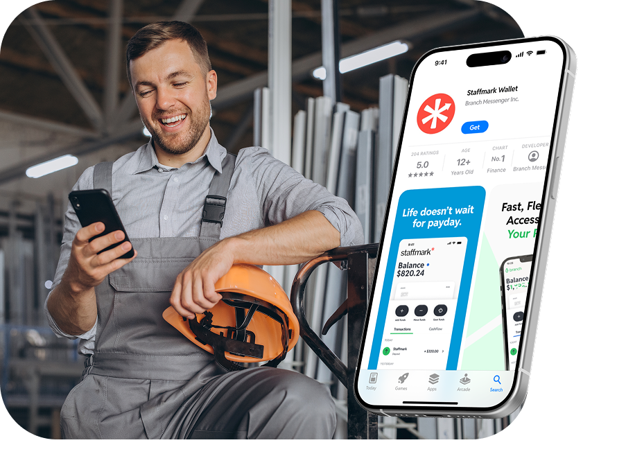 Man in work overalls holding orange helmet smiling while using a smartphone in an industrial setting.
