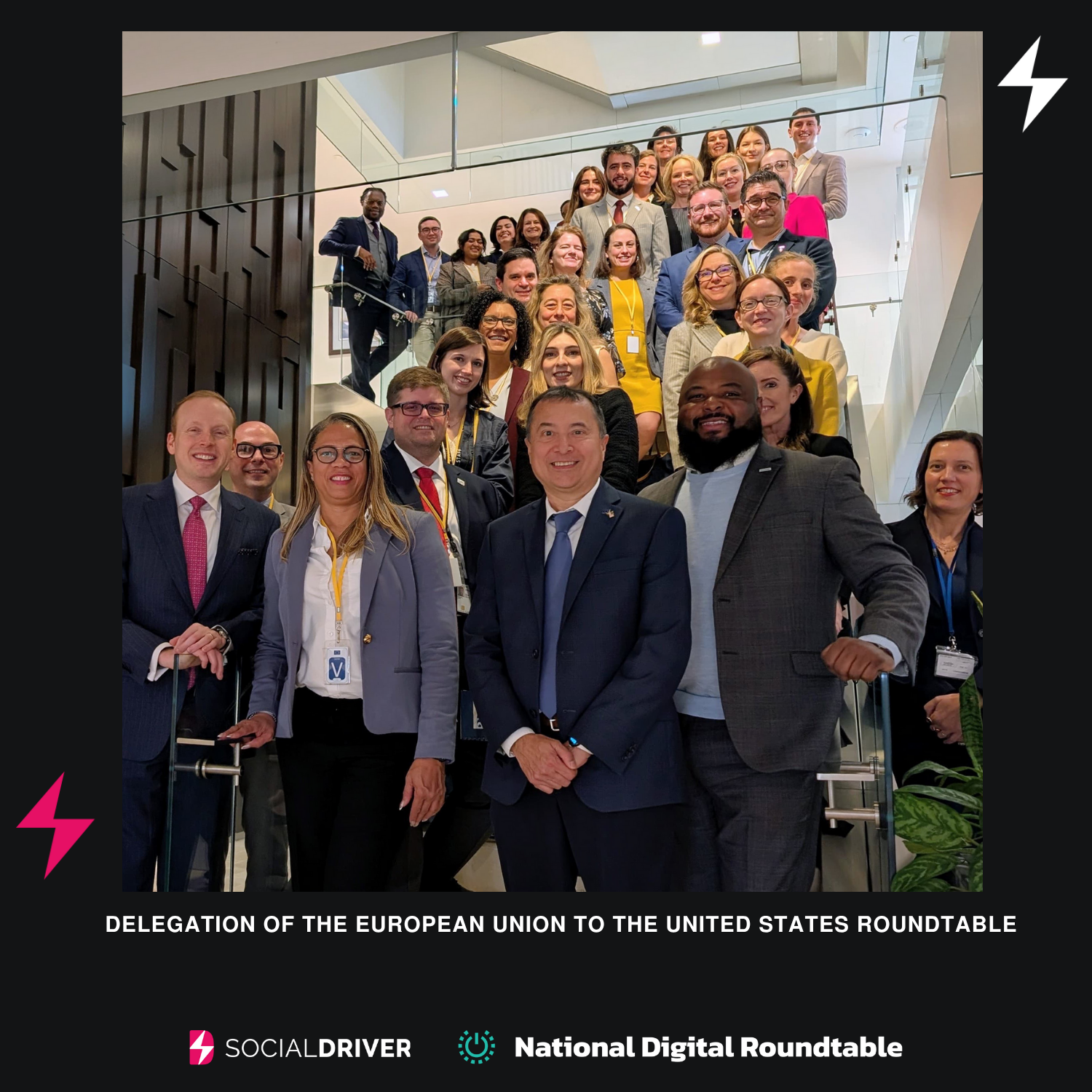 A large group of professionals stands together on a modern staircase, smiling for a group photo during a Delegation of the European Union to the United States roundtable event. The background shows bright indoor lighting and glass railings. At the bottom of the image are the Social Driver and National Digital Roundtable logos.