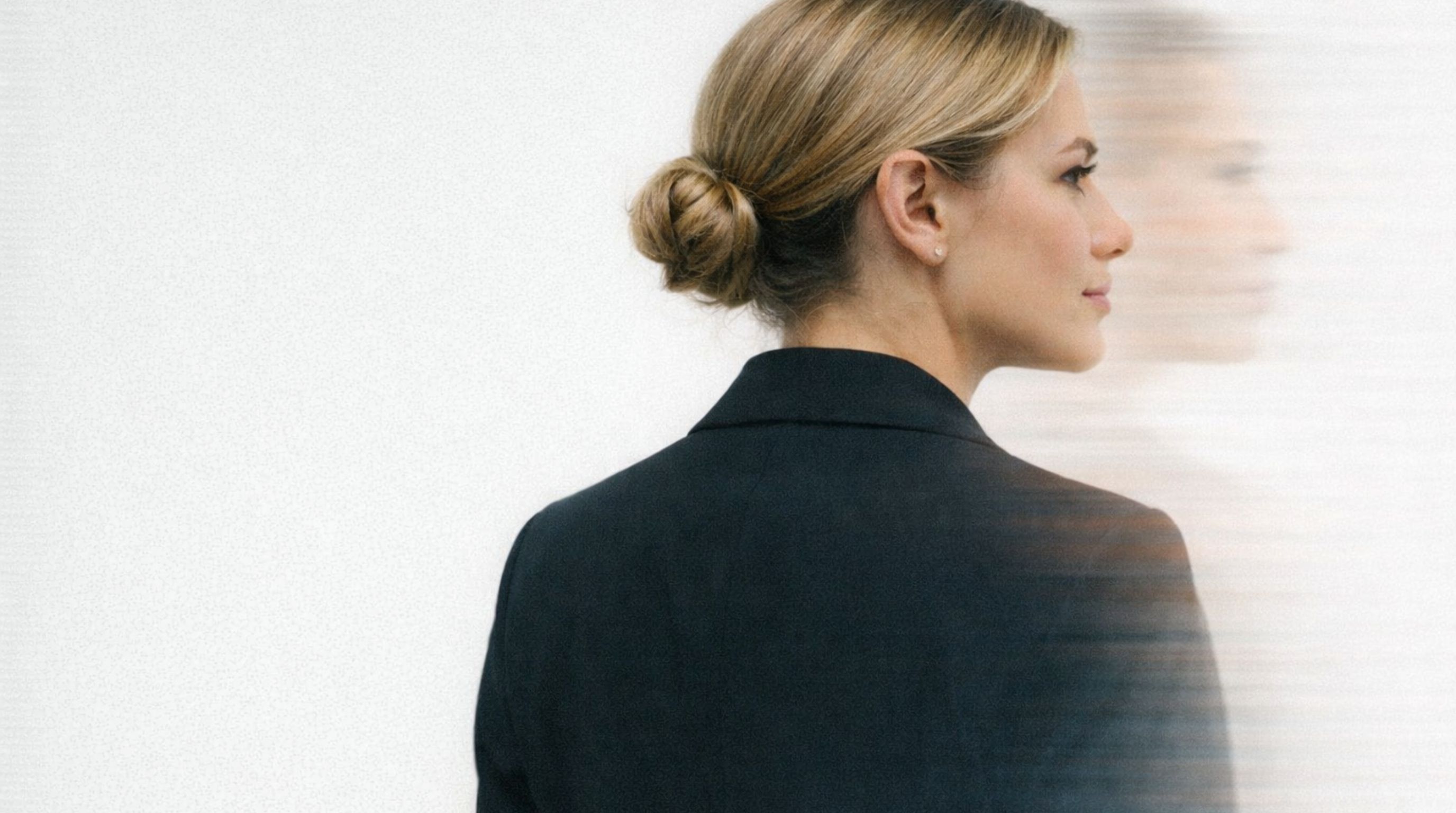 Profile of a white woman with blonde hair in a bun wearing a black blazer, with a blurred double exposure effect to her right.