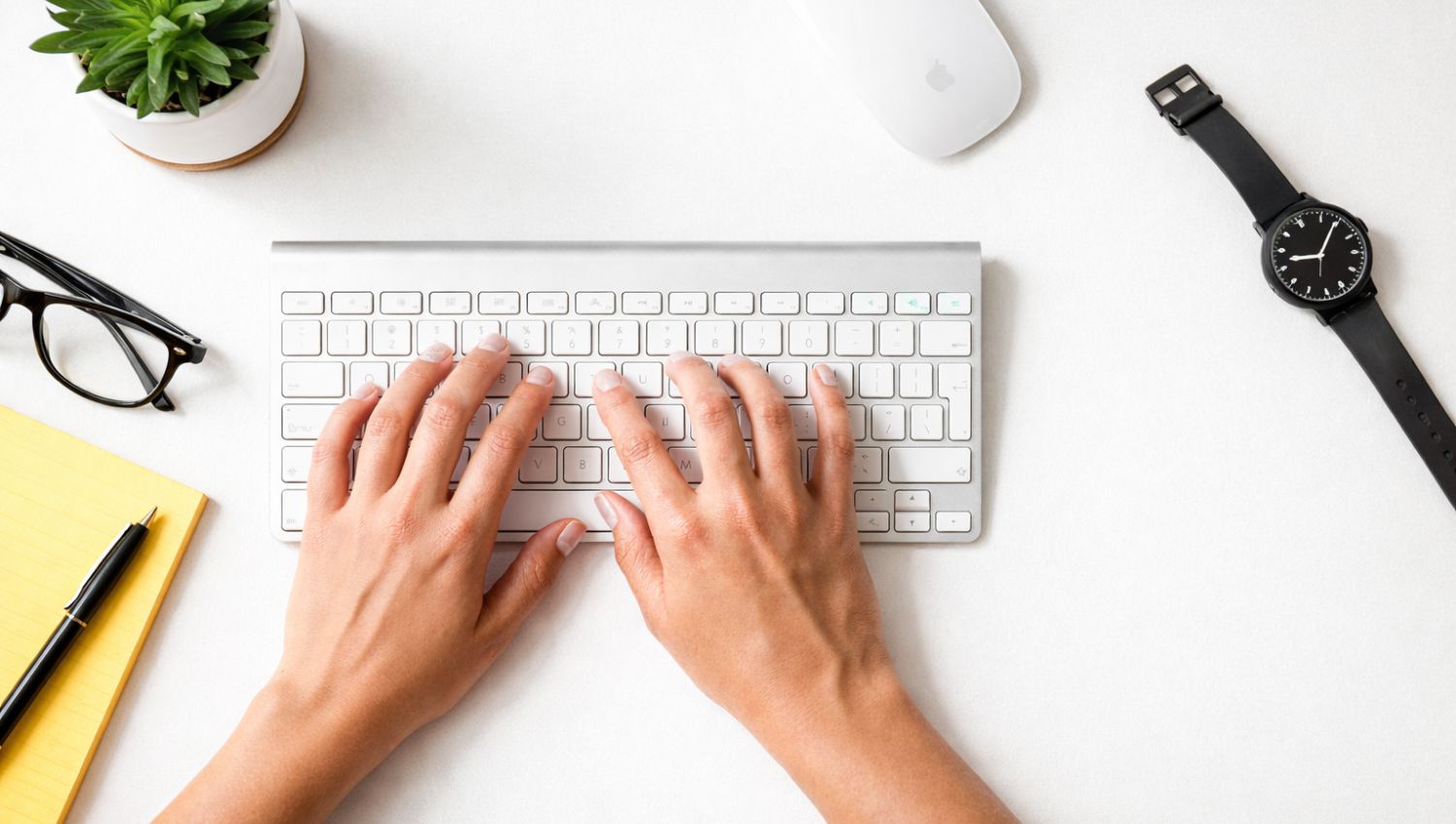 Close-up of hands typing on a keyboard at a minimal desk setup with a notebook, plant, glasses and black watch.