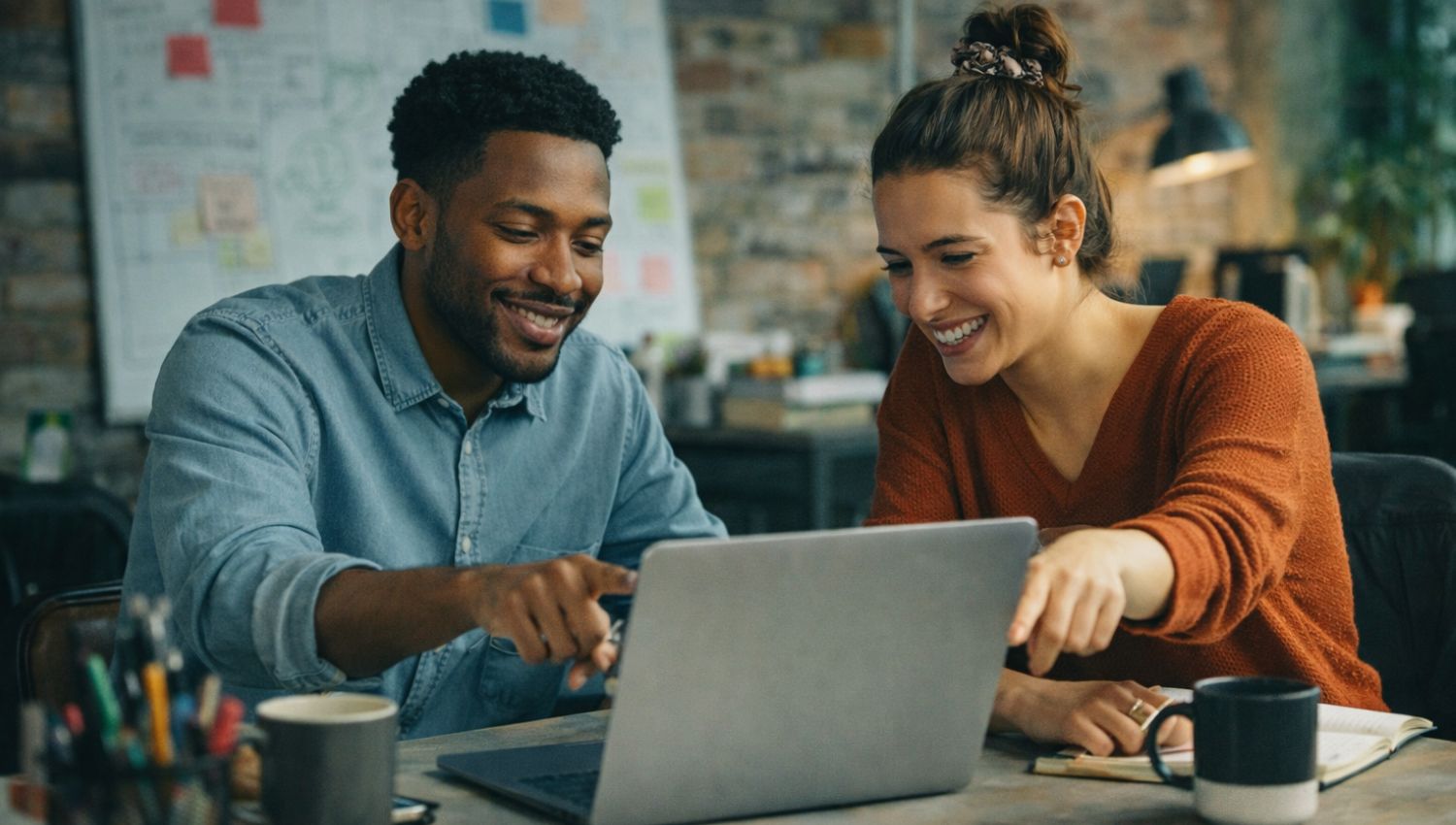 Two colleagues collaborating on a laptop at a desk in a modern creative office with notes and workspace items.