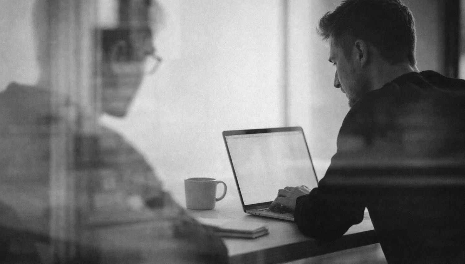 Black and white image of two people working on laptops at a desk, viewed through glass in a modern office.