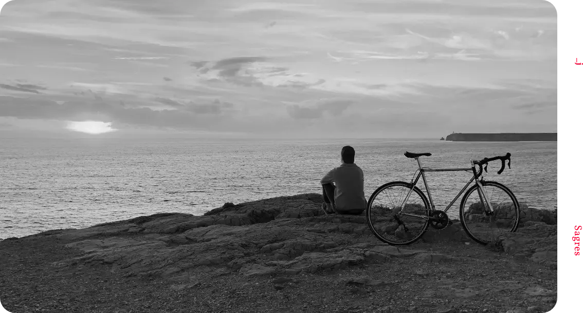 A black & white photograph of a designer sitting on a cliff overlooking a beautiful sunset coastline