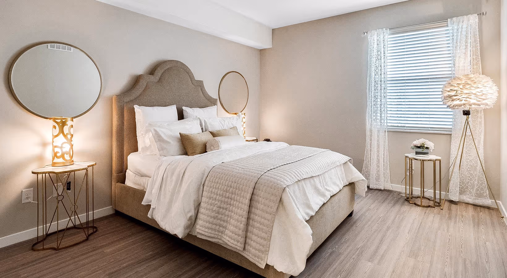 Modern bedroom with beige upholstered bed, white bedding, two round mirrors above nightstands, and a window with sheer curtains and a feathered floor lamp.