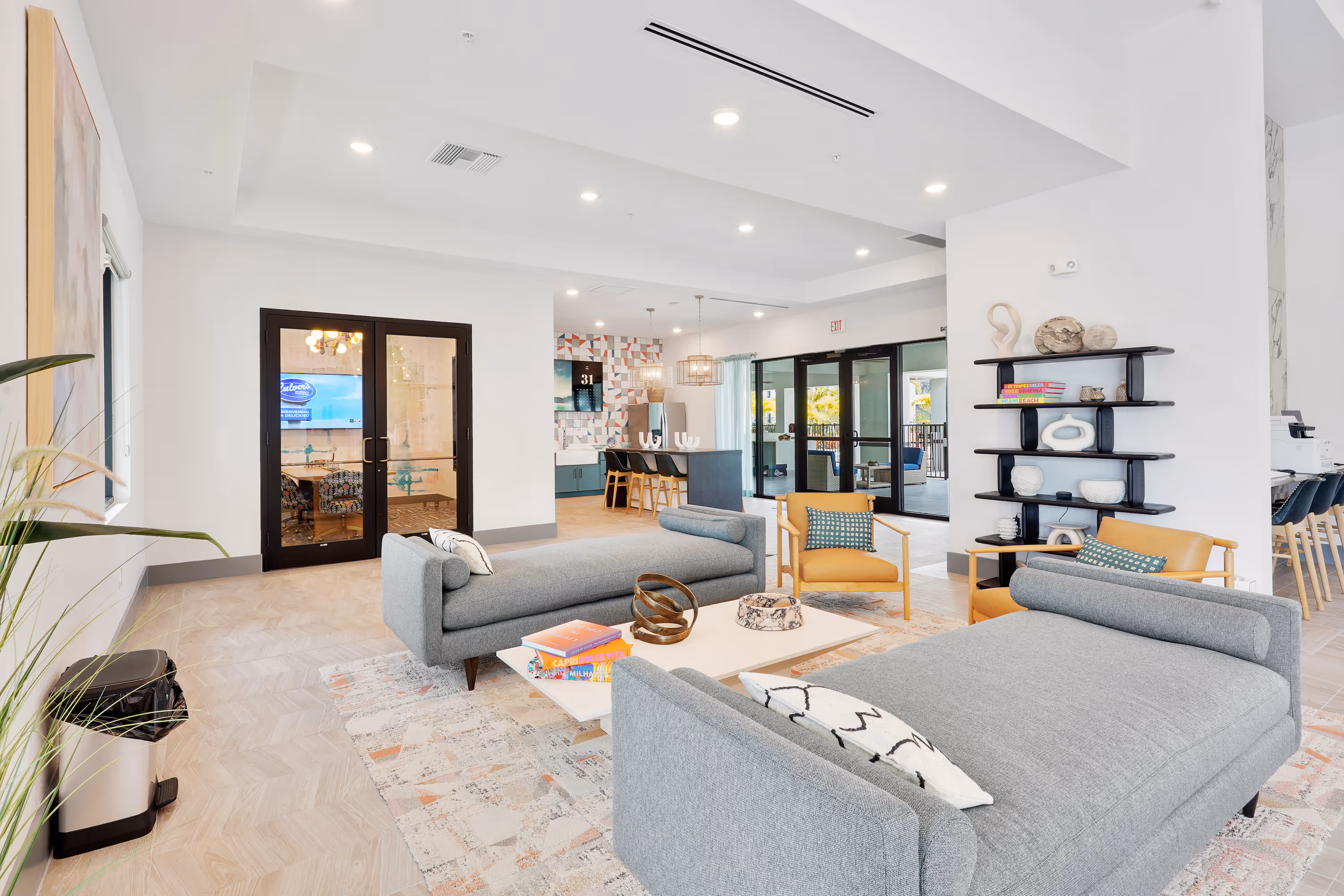 Resident clubhouse lounge area with grey couches and a black decorative shelf, showing a view into the community kitchen and business center at The Edge at Fort Myers, FL.