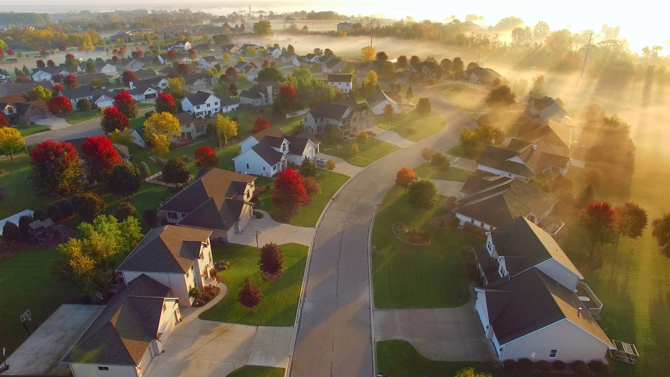 Aerial view of a suburban neighborhood with houses, colorful autumn trees, and soft morning sunlight filtering through mist.