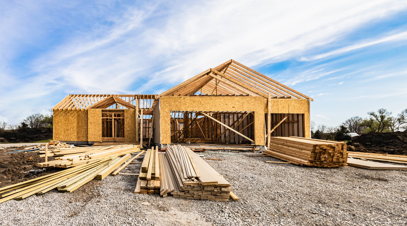 Wooden framework of a house under construction with stacks of lumber in front on a gravel lot.