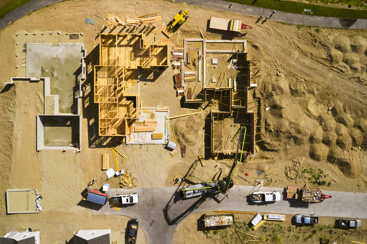 Aerial view of residential construction site showing house frameworks, construction vehicles, and materials on bare soil.