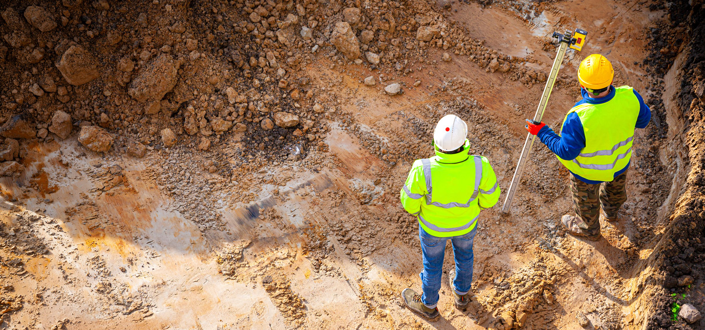 Wooden framework of a house under construction with stacks of lumber in front on a gravel lot.