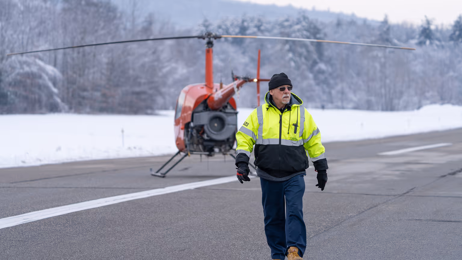 Ground crew member in high-visibility jacket walking on runway with RPX autonomous helicopter landed in background during winter operations
