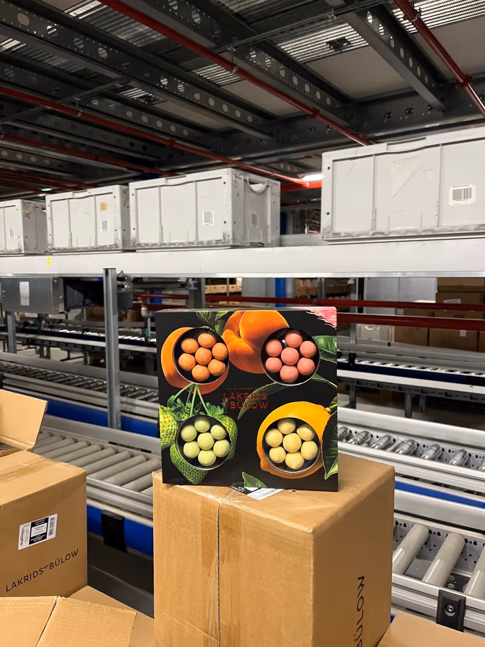 Eine bunte Lakritsbox von Lakrids by Bülow steht auf einem Karton in einem Lager mit Förderbändern und weißen Kunststoffkisten im Hintergrund.