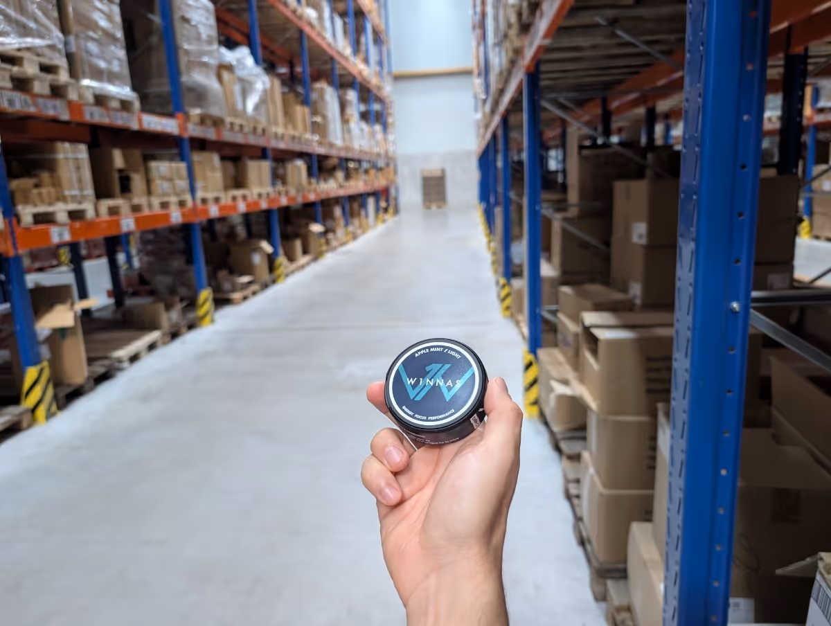 Hand hält eine Dose WINNAS Apple Mint / Light in einem Lager mit gestapelten Kartons und Regalen.