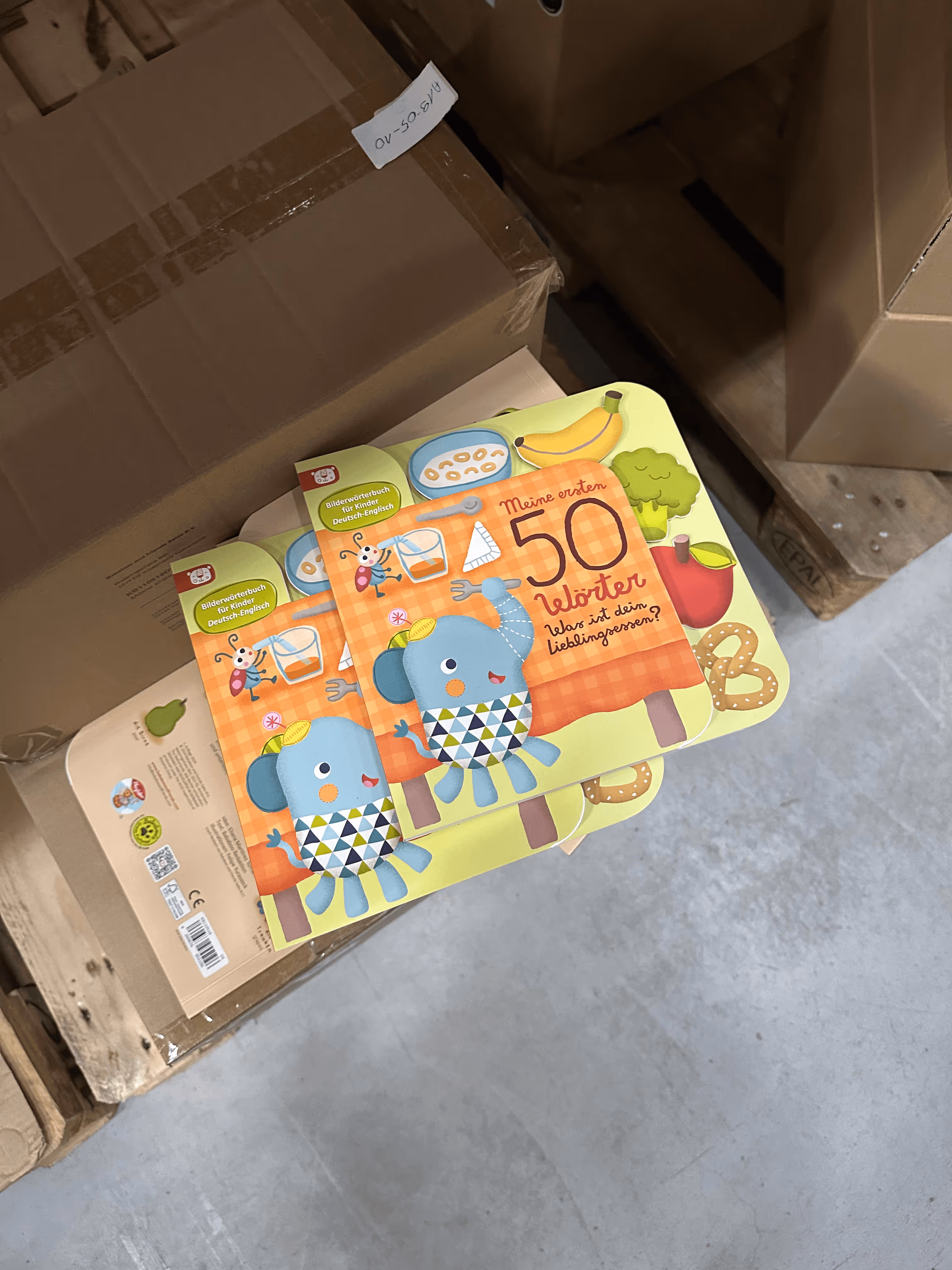 Libro per bambini «Le mie prime 50 parole» con un elefante blu e illustrazioni di cibo su una pila di imballaggi in un magazzino.