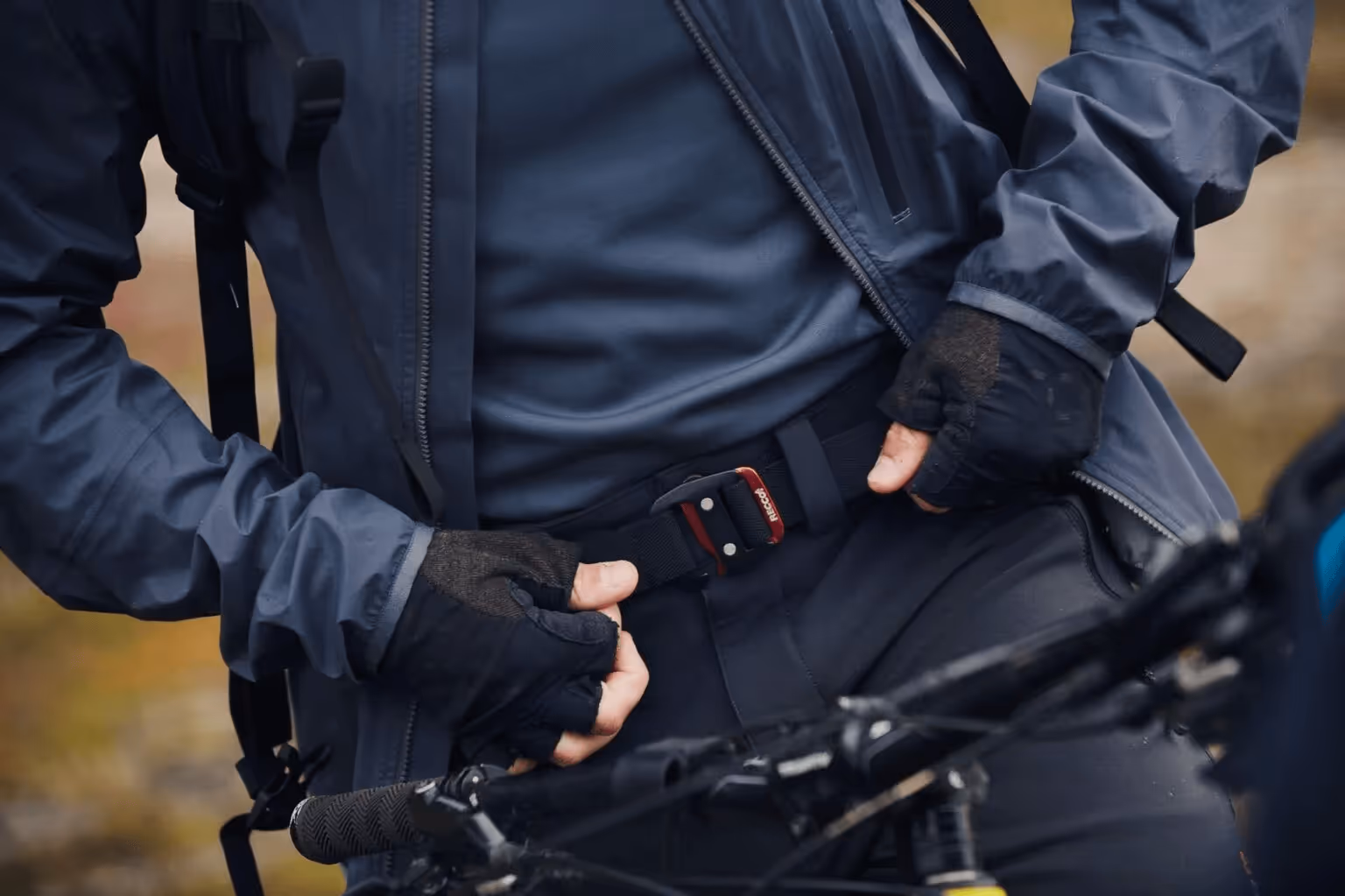 Une personne portant une veste d'extérieur bleue attache une ceinture RECCO noire à la zone de la taille, devant le guidon de vélo.