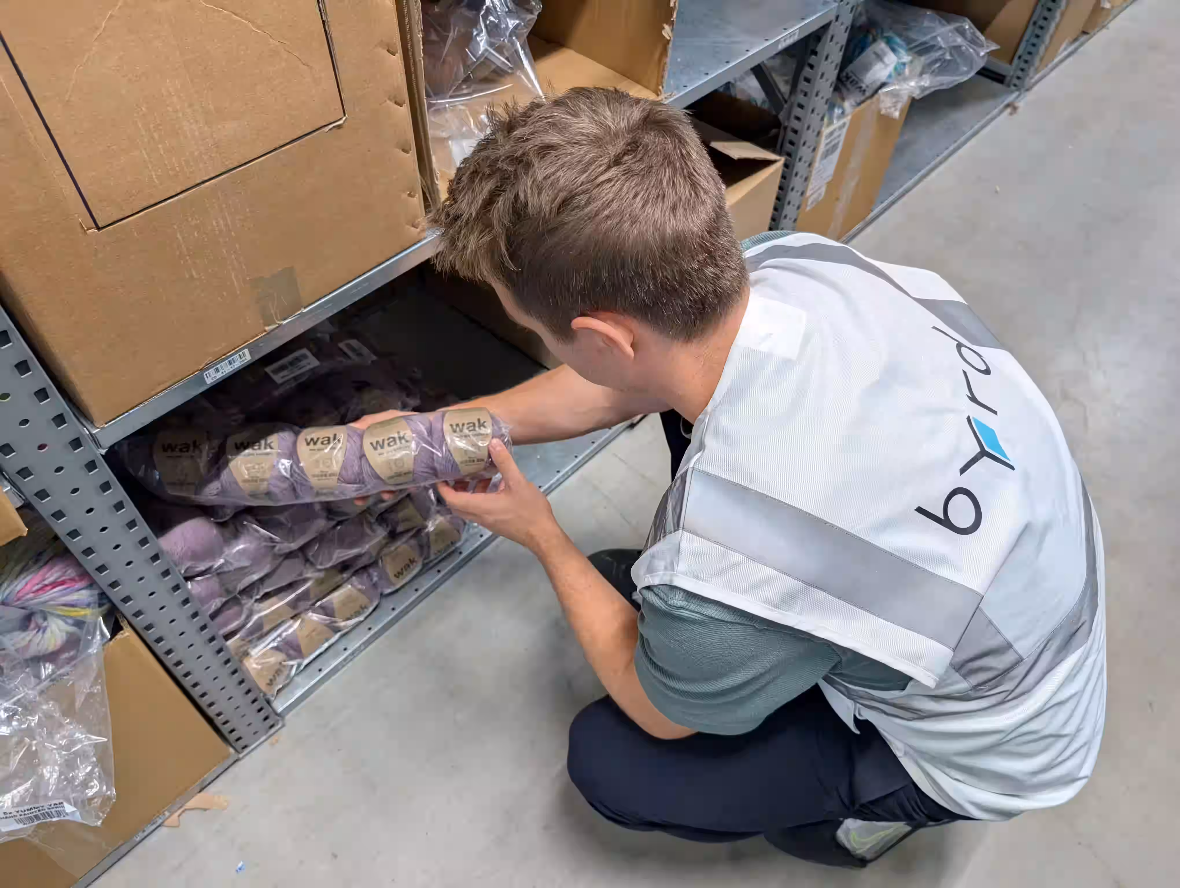 Un homme vêtu d'un gilet portant l'inscription « byrd » s'agenouille devant une étagère et tient un paquet de laine tricotée portant la marque « We are Knitters ».