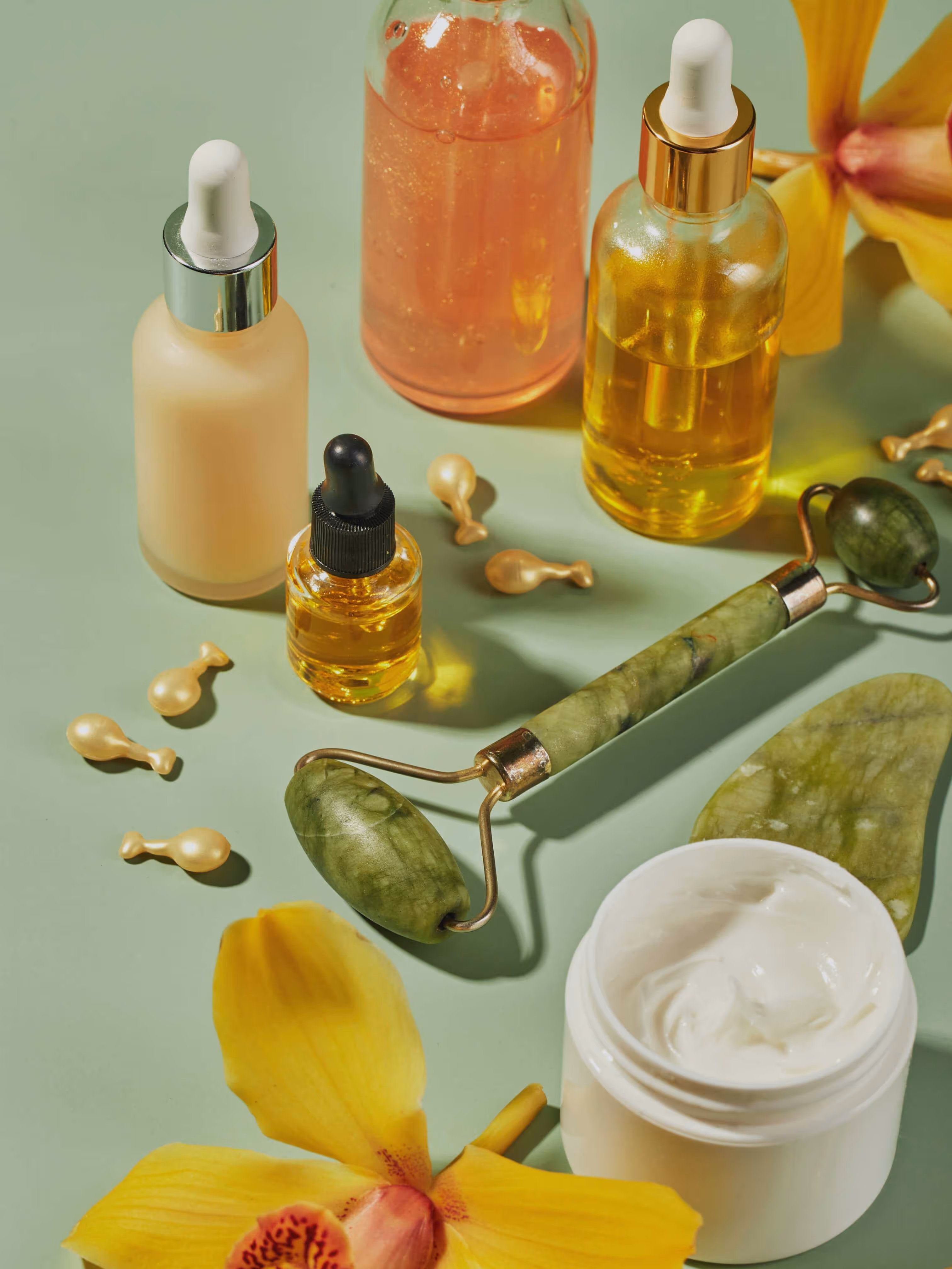 Diversos productos para el cuidado de la piel con masajeadores faciales de jade y flores de orquídeas amarillas sobre un fondo verde.