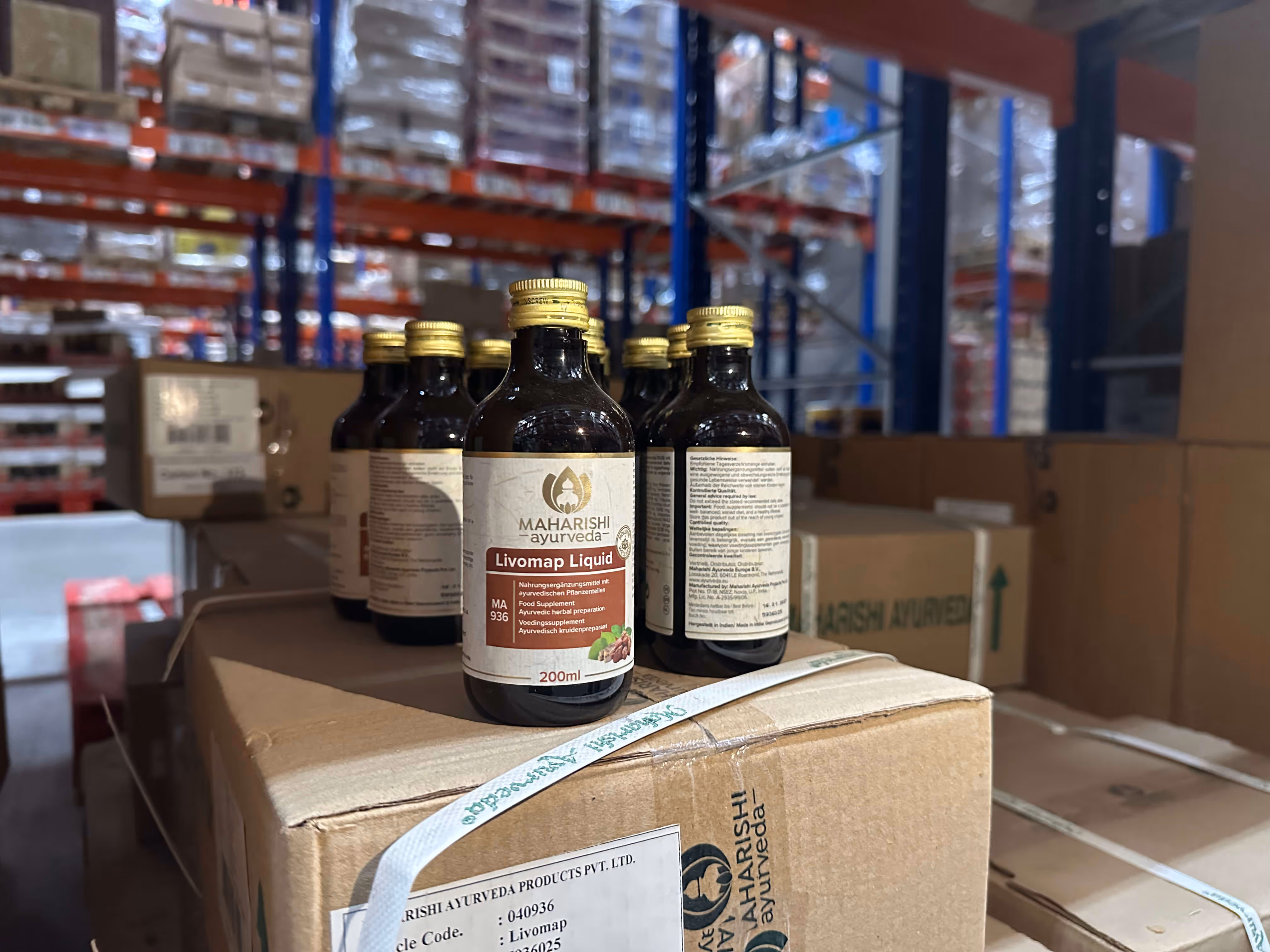 Il y a plusieurs bouteilles de suppléments nutritionnels Maharishi Ayurveda Livomap Liquid dans une boîte en carton dans une salle de stockage.