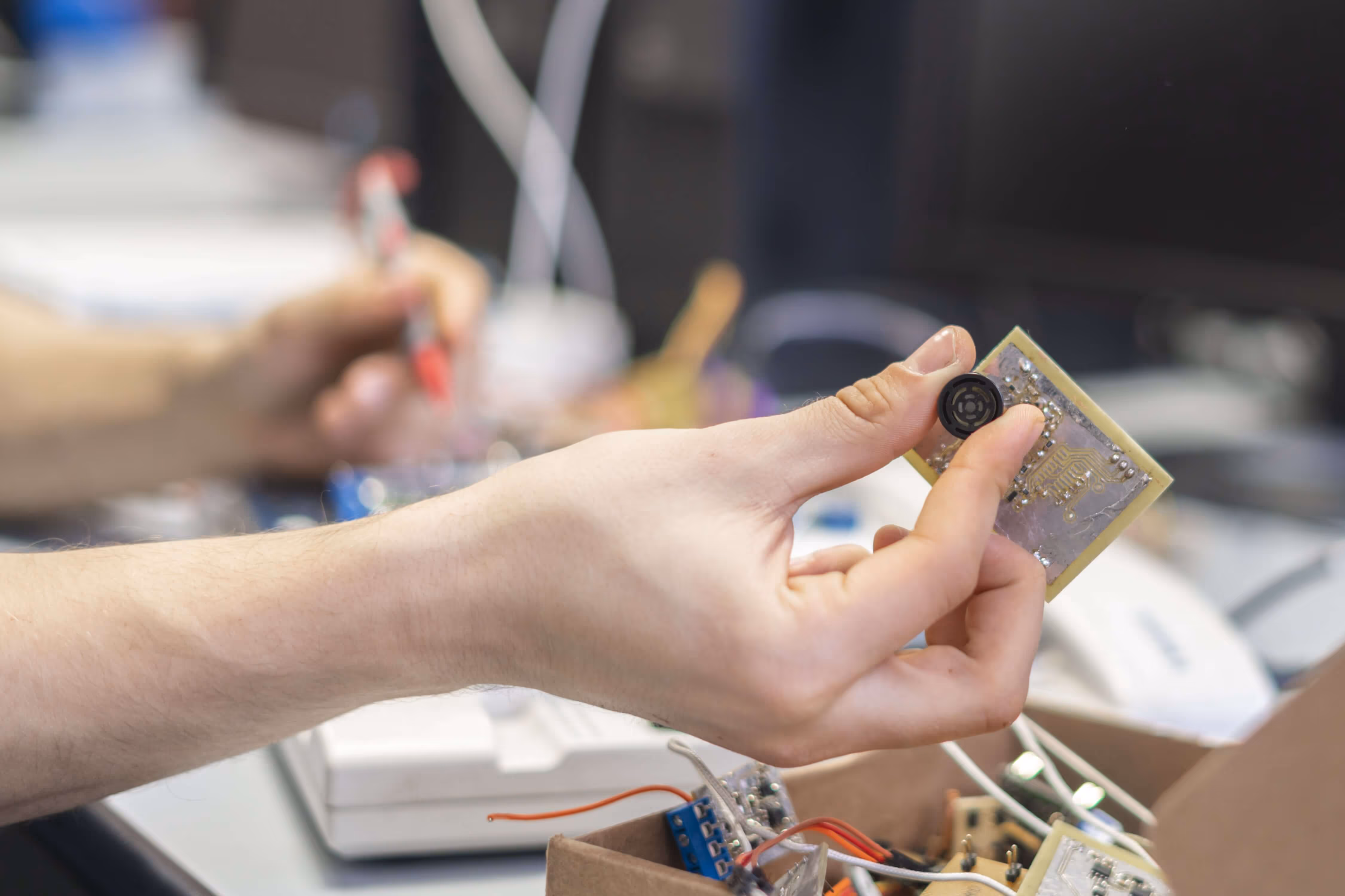 La mano tiene in mano un circuito stampato con un piccolo componente nero in un'area di lavoro elettronica.