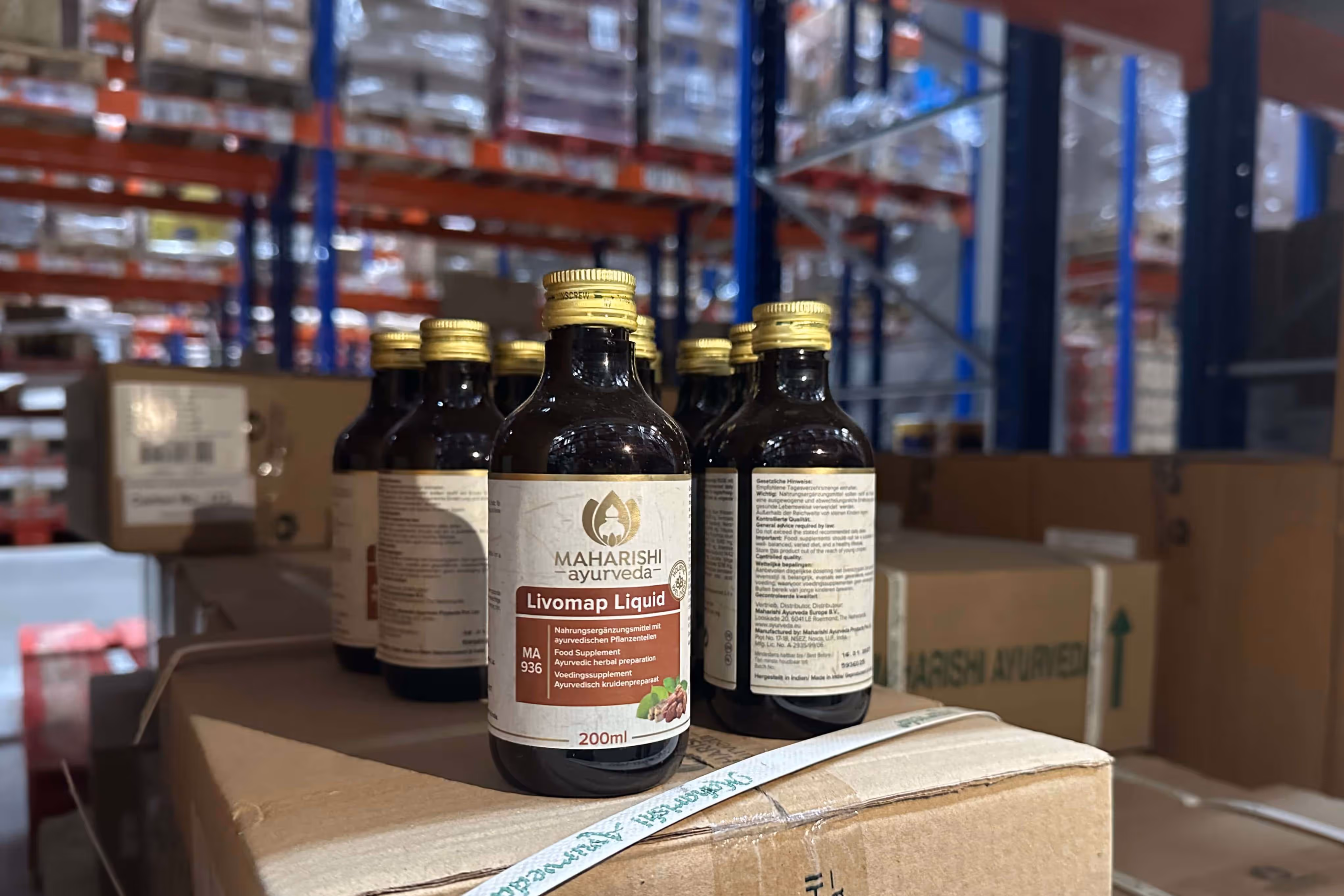 There are several bottles of Maharishi Ayurveda Livomap Liquid nutritional supplements on a cardboard box in a warehouse.