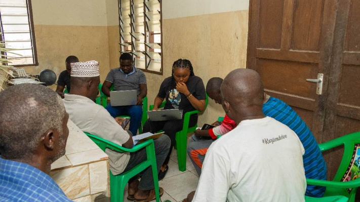 The image shows a group of people gathered around a table, each engaged in working on laptops, with a wall featuring a tiled and wooden wall, and some individuals wearing traditional attire.AI-generated content may be incorrect.