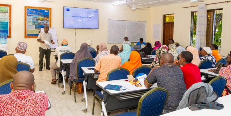 The image shows a classroom setting with a whiteboard and projector, where a person is presenting to a group of people seated in rows, some of them wearing colorful clothing.AI-generated content may be incorrect.