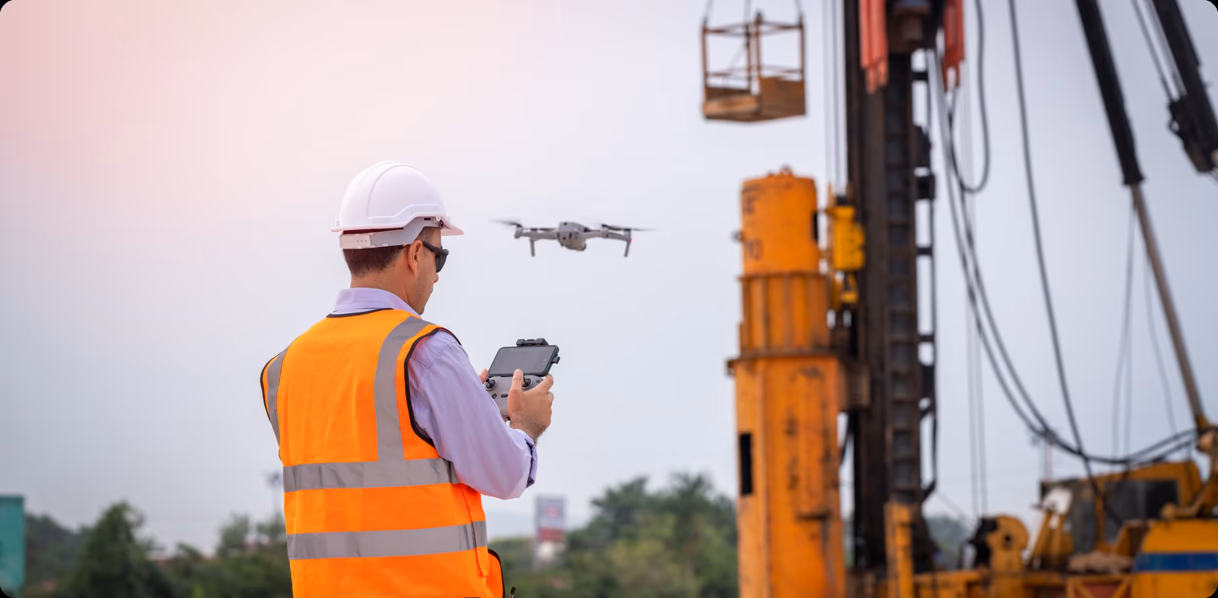 Construction worker in a high-visibility vest and hard hat piloting a drone near heavy industrial equipment, demonstrating on-site drone operations for infrastructure inspection and safety monitoring.