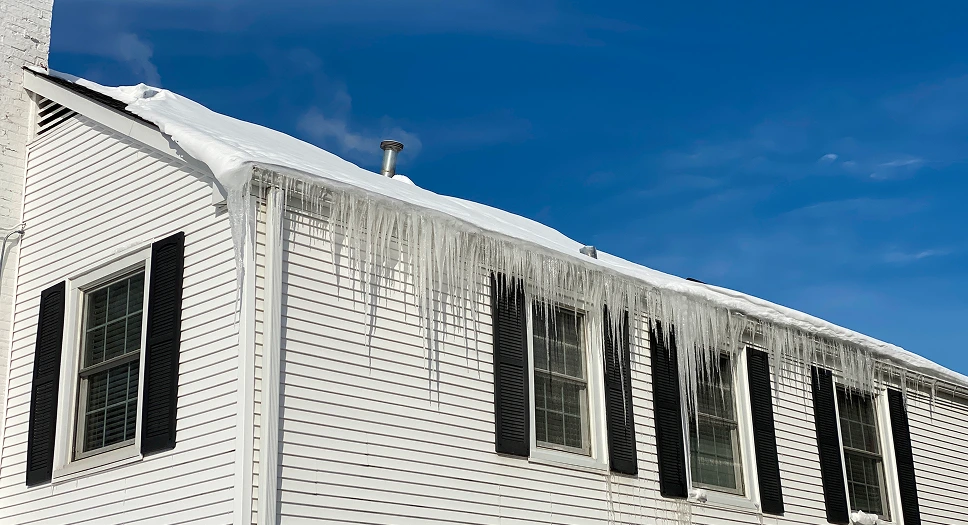 Ice dams and icicles hanging from a snowy roof, showing winter roof leak risks caused by ice buildup