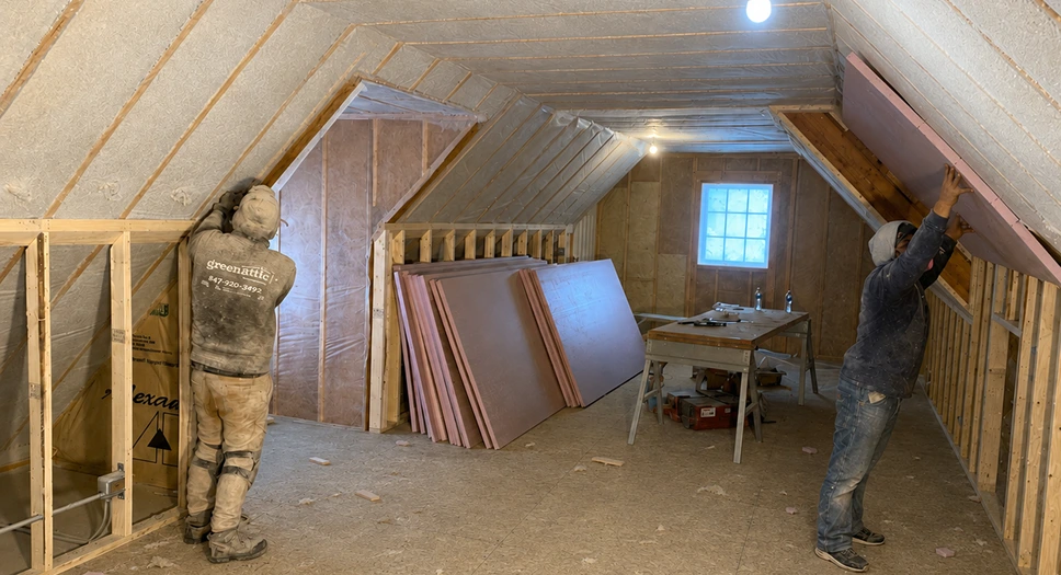 Workers installing insulation and soundproofing materials in an attic remodel with exposed framing and spray foam insulation.