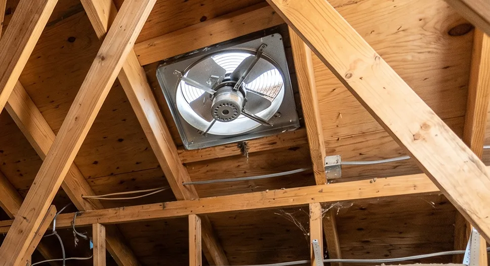 Attic fan installed in the roof deck between wood rafters for attic ventilation.