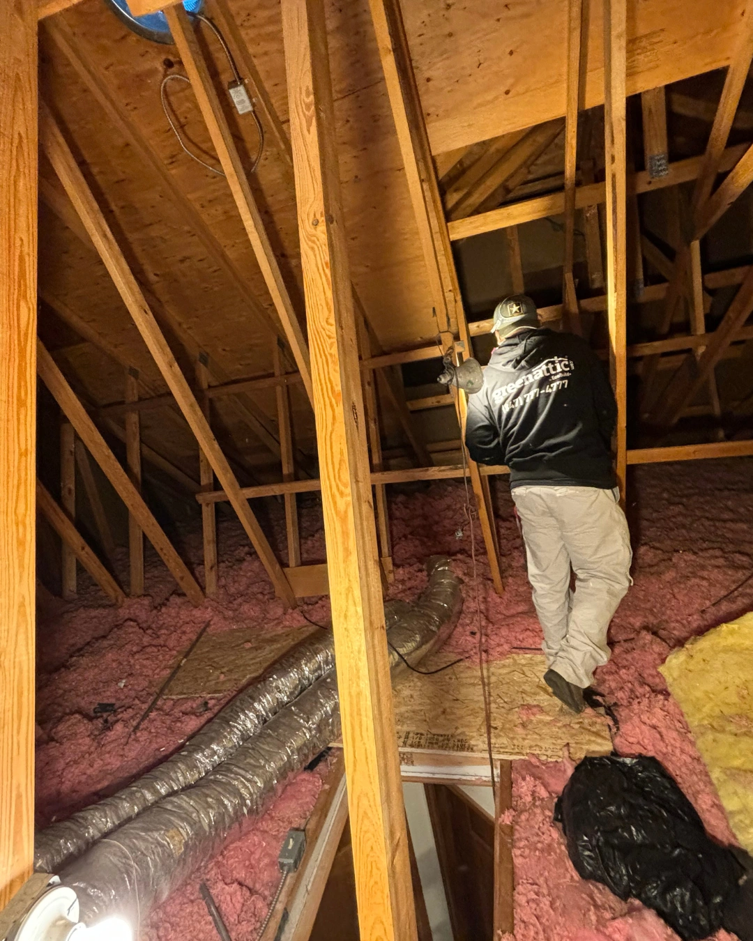 Green Attic technician in a branded jacket moving along plank boards in an attic with pink fiberglass insulation and flexible ductwork