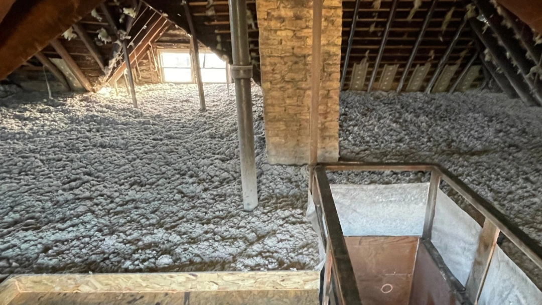 Attic floor fully covered with blown-in cellulose insulation around a brick chimney, with a railed access opening in the foreground and a gable window at the far end