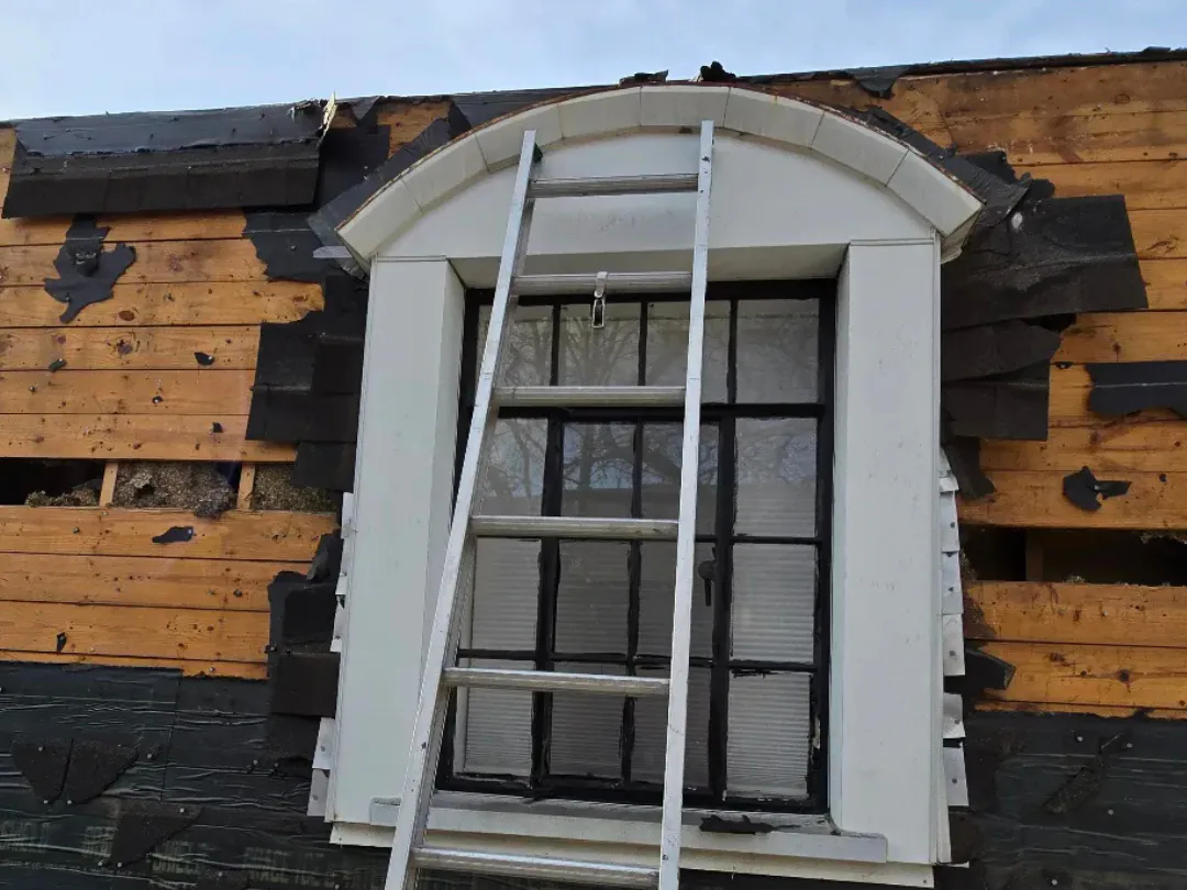 Roof tear-off in progress with shingles stripped back to bare boards and underlayment exposed above a window