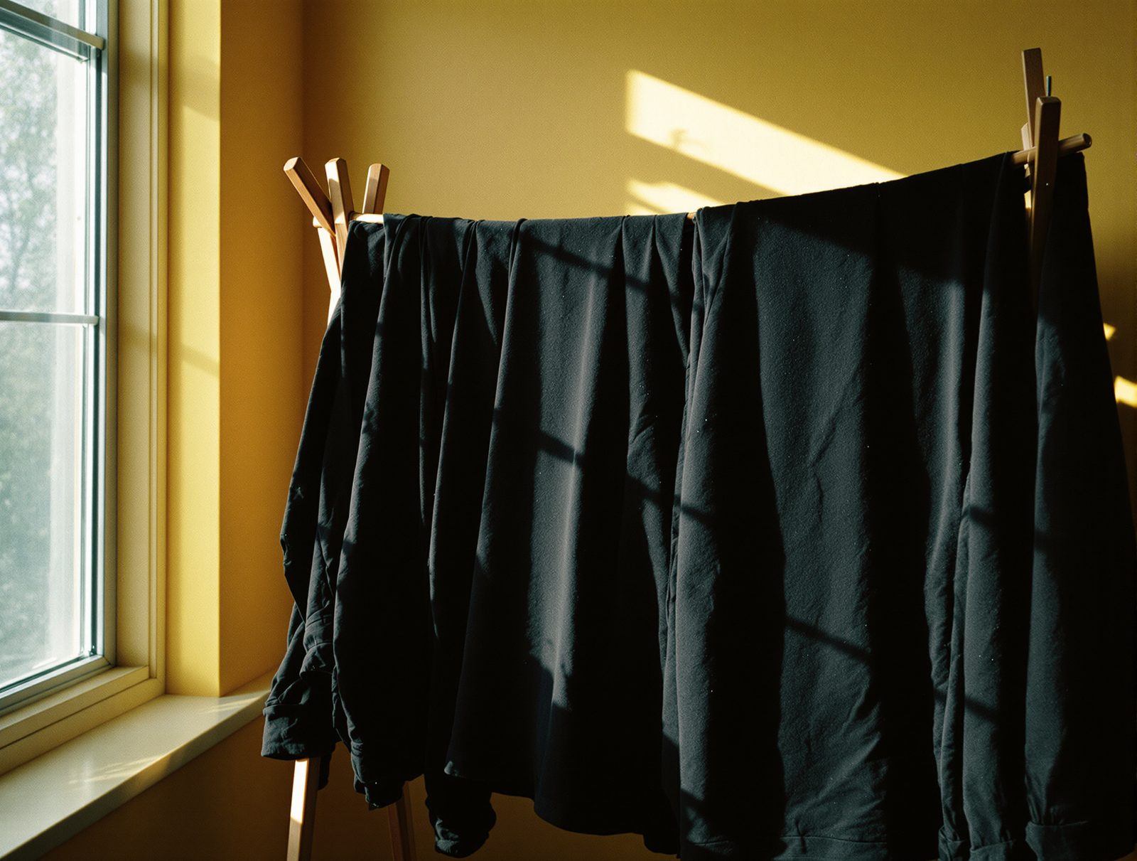 Vêtements noirs suspendus sur un séchoir en bois à ombre intérieure, lumière naturelle douce, ambiance vintage