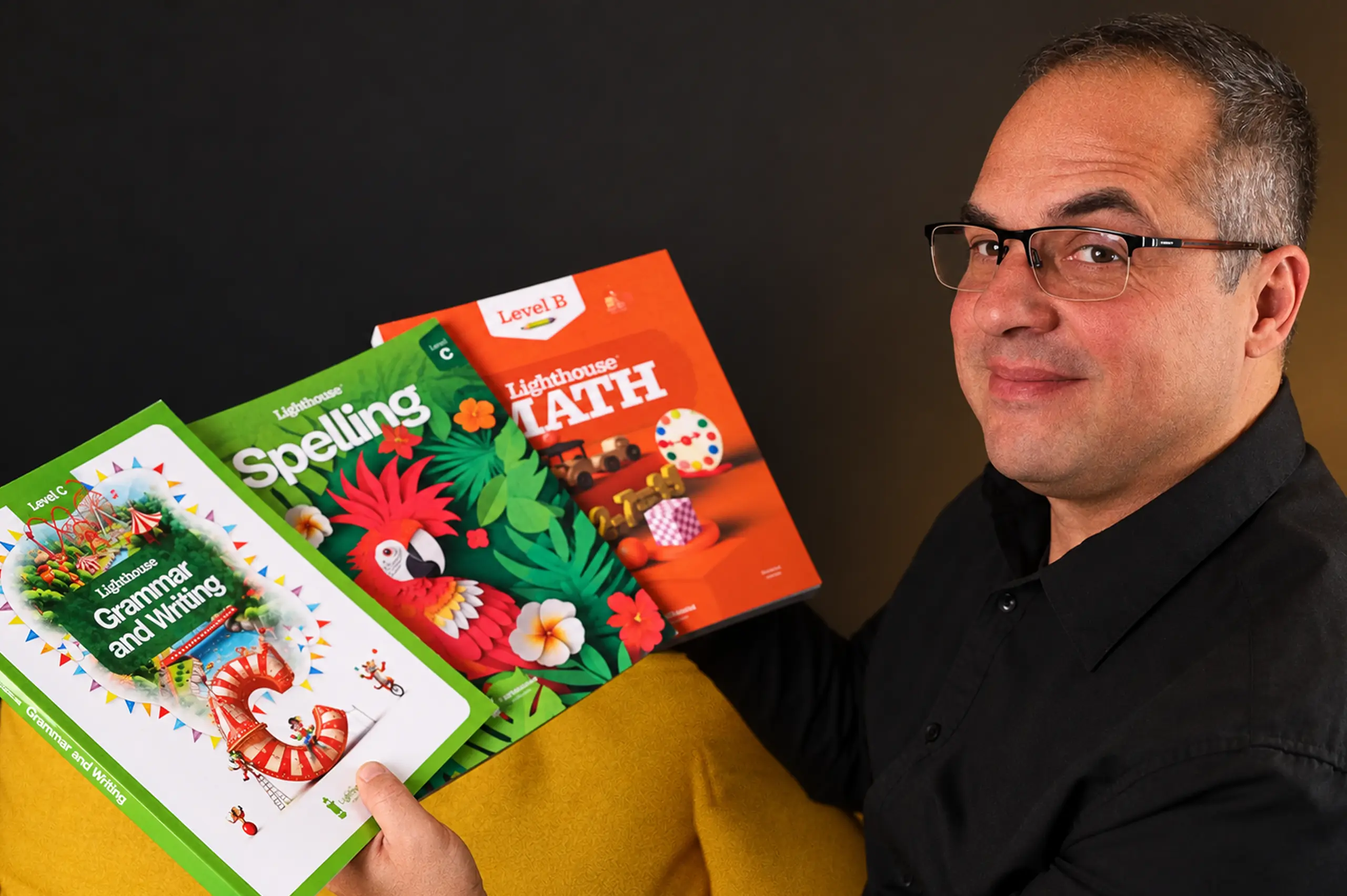 Man wearing glasses and a black shirt holding three colorful textbooks on grammar, spelling, and math.