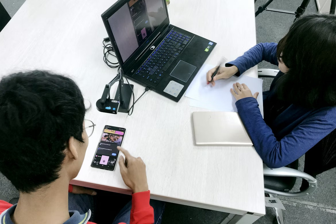 Two people participating in an in-person user testing session. One person uses a smartphone to complete tasks while the other takes notes beside a laptop capturing the session.